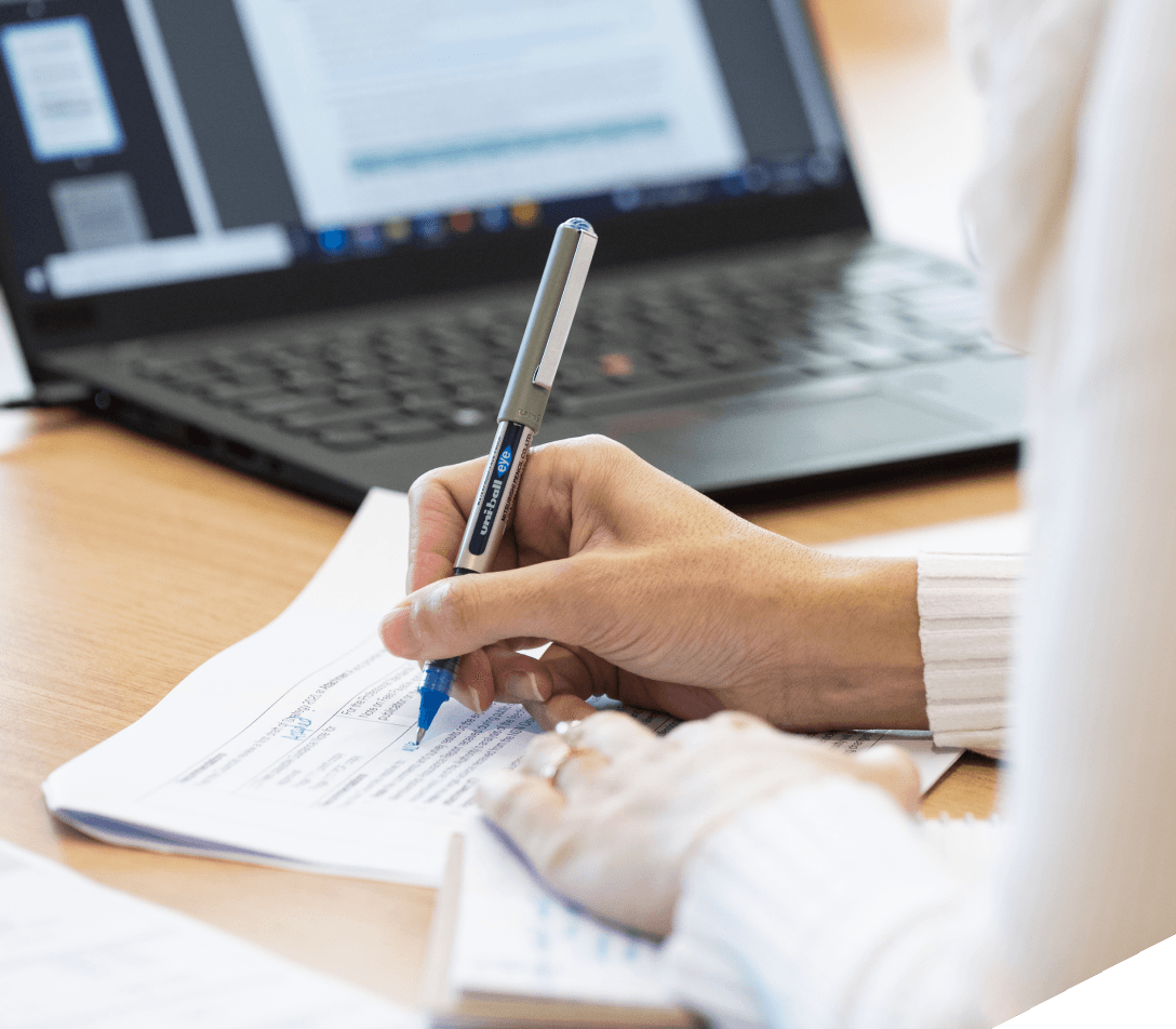 Close up of hand holding a pen and writing on paper with laptop in the background